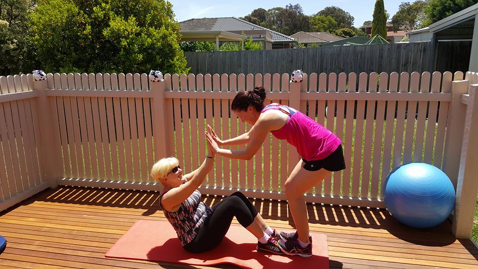A one on one PT session outdoors. A female client doing a sit-up and clapping hands with Erin, the trainer.